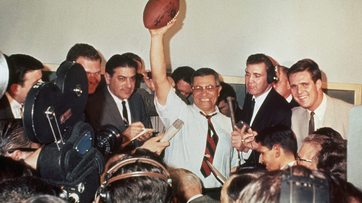 Green Bay Packers coach Vince Lombardi raises a football in victory, surrounded by reporters covering the first Super Bowl in 1967.
